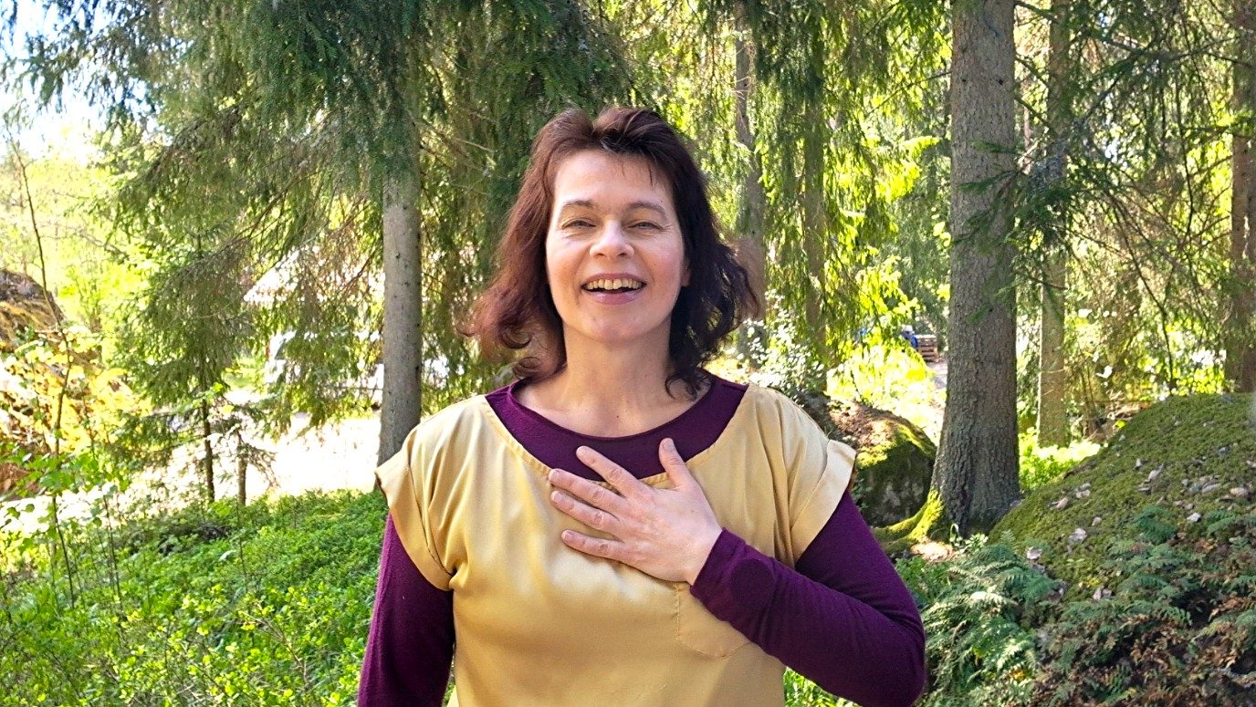 Satu Haapala Resonoiva Ääni - kuvassa hymyilevänä käsi rintakehän päällä, taustalla metsäistä luontoa